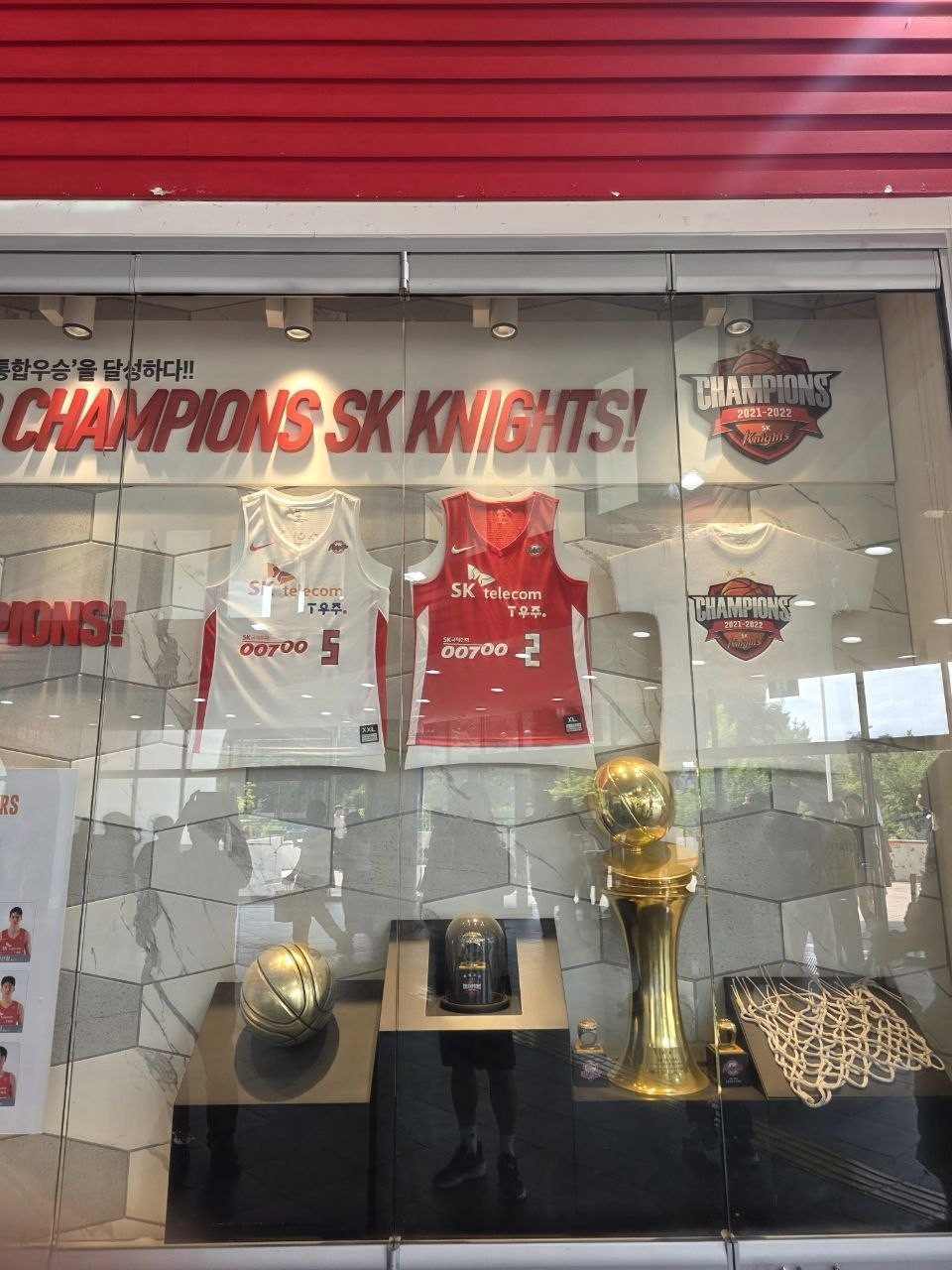 체육관 입구 유리 진열장 — CHAMPIONS SK KNIGHTS 문구와 우승 기념 유니폼, 금색 트로피, 농구공, 우승 그물이 전시된 모습