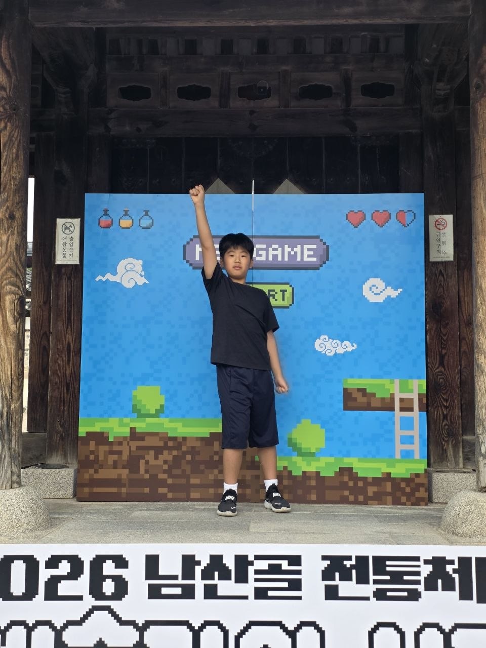 남산골한옥마을 한옥 대문 아래 옛날 게임 화면처럼 꾸민 NEW GAME 포토존 앞에서 아이가 주먹을 들어 올린 모습과 2026 남산골 전통체험 예술가의 시간 현수막