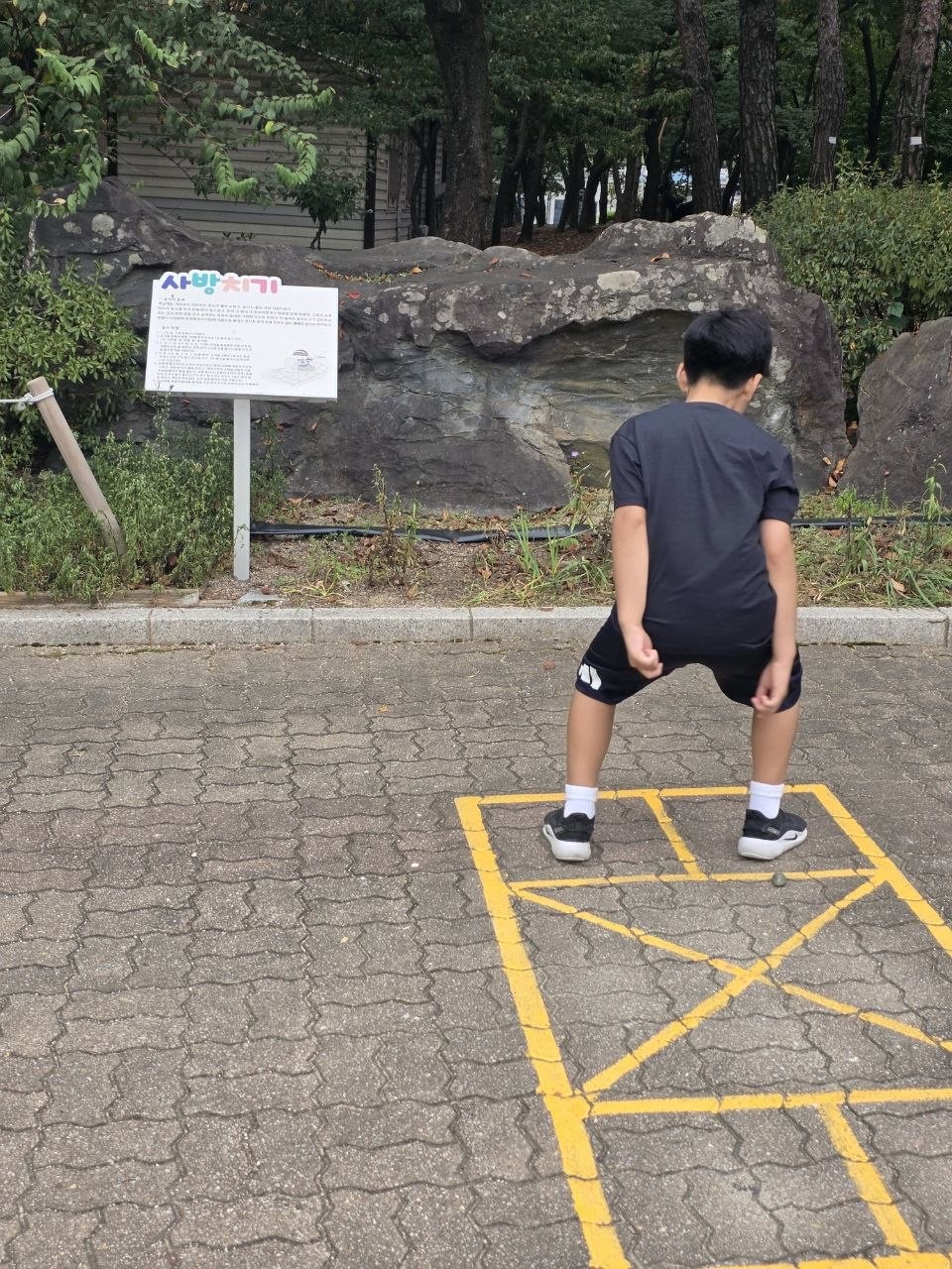 한옥마을 옆 보도블록 바닥에 노란 선으로 그려진 사방치기 판에서 아이가 뛰려고 자세를 잡은 뒷모습, 뒤에 사방치기 규칙 안내판