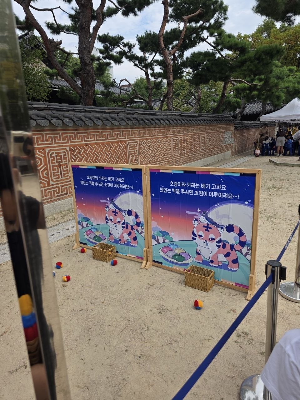 운현궁 담장 앞에 호랑이와 까치 그림판 두 개가 서 있고 그 앞 바구니 주변에 떡 모양 콩주머니가 흩어져 있는 호랑이 떡 먹이기 놀이판