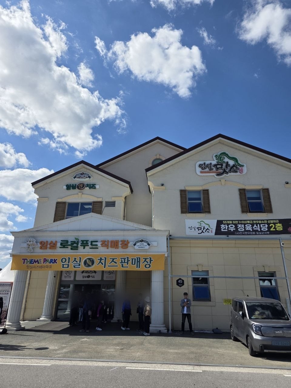 임실치즈테마파크 주차장 앞 임실 로컬푸드 직매장과 임실N치즈판매장 건물 — 2층 한우 정육식당 현수막이 걸려 있고 입구 앞에 사람들이 줄 서 있는 모습